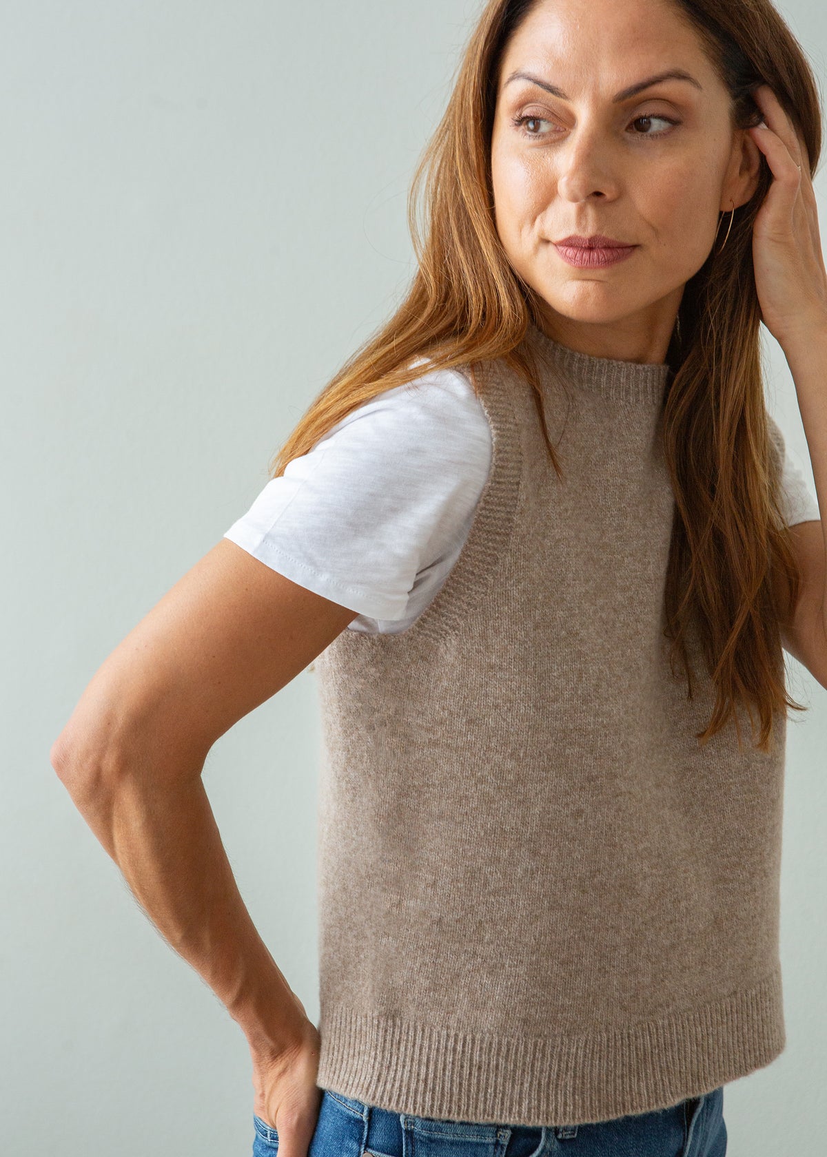 A woman wearing a sleeveless taupe knit sweater vest over a white t-shirt and blue jeans, posing against a light gray background.