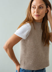 A woman wearing a sleeveless taupe knit sweater vest over a white t-shirt and blue jeans, posing against a light gray background.