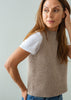 A woman wearing a sleeveless taupe knit sweater vest over a white t-shirt and blue jeans, posing against a light gray background.