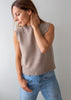 A woman wearing a sleeveless knit sweater vest in a taupe-beige color paired with blue jeans, standing against a plain light gray wall.