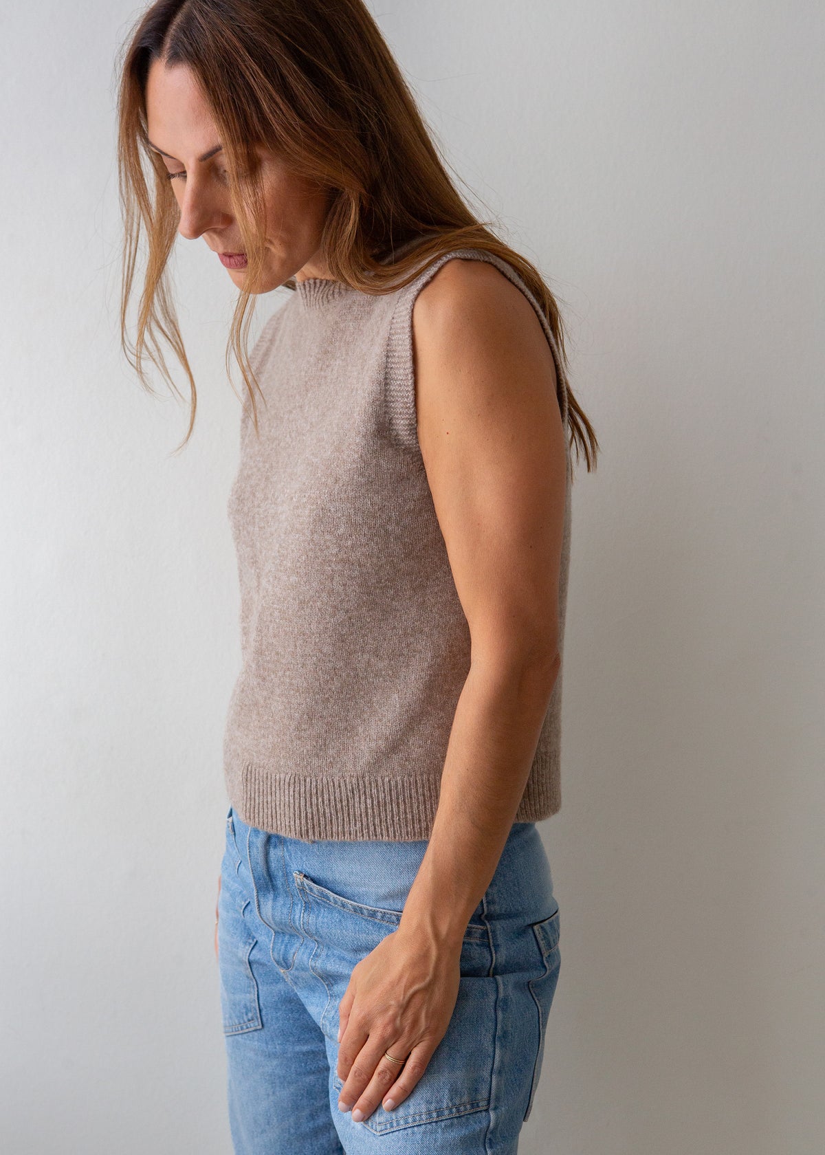 A woman wearing a sleeveless, ribbed-knit beige sweater vest and light blue jeans, standing against a plain light gray wall.