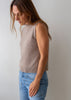 A woman wearing a sleeveless, ribbed-knit beige sweater vest and light blue jeans, standing against a plain light gray wall.