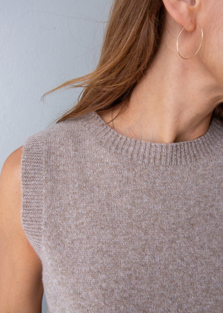 Close-up of a woman wearing a sleeveless ribbed knit sweater in a light taupe-beige color, paired with a thin gold hoop earring.