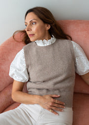 A woman wearing a taupe knit sweater vest over a white eyelet puff-sleeve blouse and white trousers, seated on a dusty pink armchair against a light gray wall.