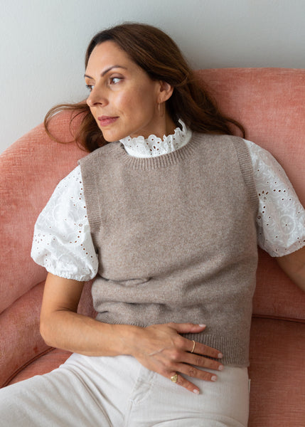 A woman wearing a taupe knit sweater vest over a white eyelet puff-sleeve blouse and white trousers, seated on a dusty pink armchair against a light gray wall.