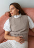 A woman wearing a taupe knit sweater vest over a white eyelet puff-sleeve blouse and white trousers, seated on a dusty pink armchair against a light gray wall.