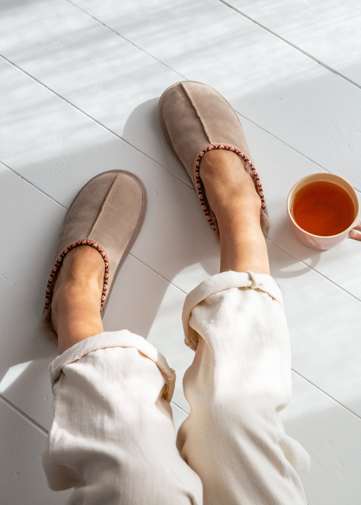 Person wearing beige sheepskin slippers on a white tiled floor with a cup of tea nearby.