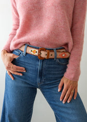 A close-up of a person wearing a pink knit sweater, blue jeans, and a decorative Guatemalan belt with a brown leather buckle. The belt has a tan base with colorful geometric patterns in orange, yellow, and white.
