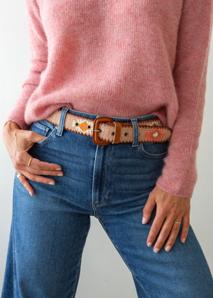 A close-up of a person wearing a pink knit sweater, blue jeans, and a decorative Guatemalan belt with a brown leather buckle. The belt has a tan base with colorful geometric patterns in orange, yellow, and white.