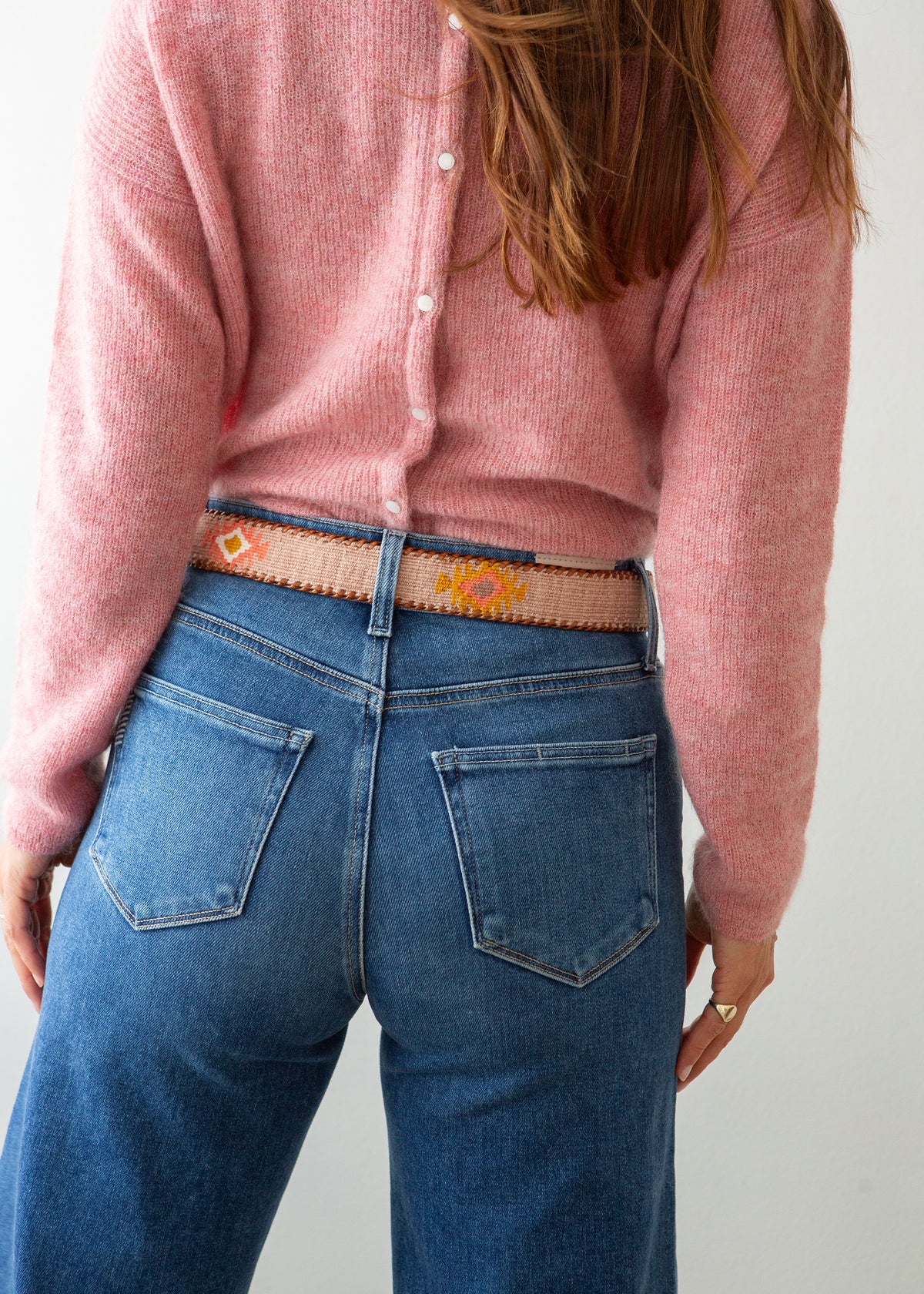 A close-up of a person wearing a pink knit sweater, blue jeans, and a decorative Guatemalan belt with a brown leather buckle. The belt has a tan base with colorful geometric patterns in orange, yellow, and white.
