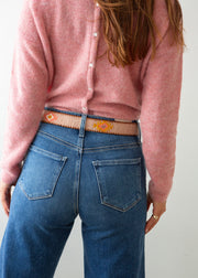 A close-up of a person wearing a pink knit sweater, blue jeans, and a decorative Guatemalan belt with a brown leather buckle. The belt has a tan base with colorful geometric patterns in orange, yellow, and white.