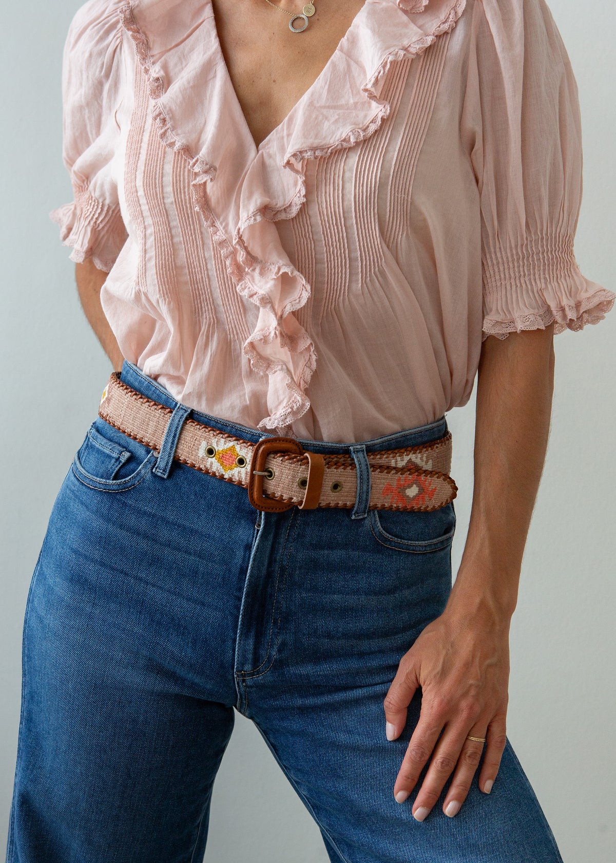 A close-up of a person wearing a pink knit sweater, blue jeans, and a decorative Guatemalan belt with a brown leather buckle. The belt has a tan base with colorful geometric patterns in orange, yellow, and white.