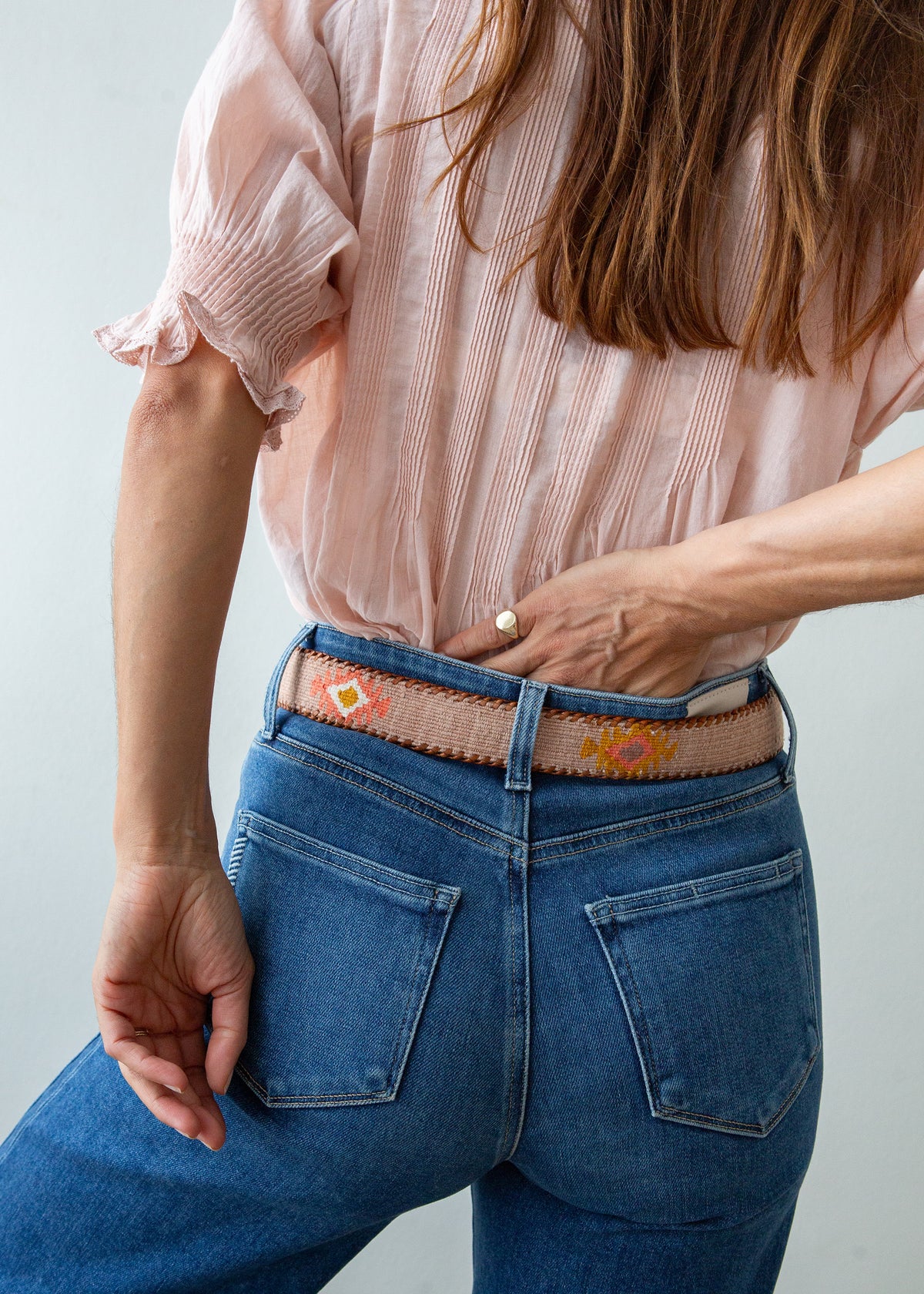 A close-up of a person wearing a pink blouse, blue jeans, and a decorative Guatemalan belt with a brown leather buckle. The belt has a tan base with colorful geometric patterns in orange, yellow, and white.