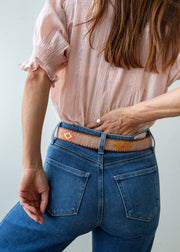 A close-up of a person wearing a pink blouse, blue jeans, and a decorative Guatemalan belt with a brown leather buckle. The belt has a tan base with colorful geometric patterns in orange, yellow, and white.
