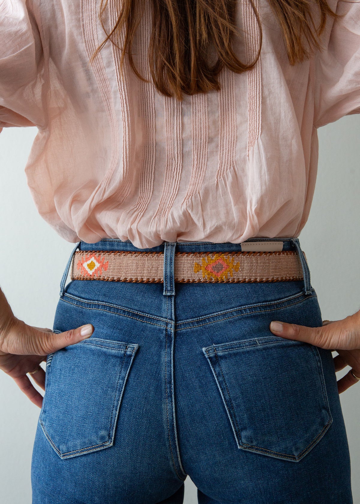 A close-up back view of a person wearing a woven belt with geometric and floral patterns in orange, yellow, and pink, paired with blue jeans and a light pink blouse.