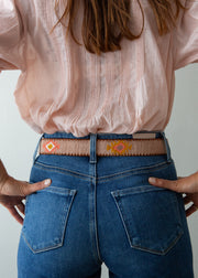 A close-up back view of a person wearing a woven belt with geometric and floral patterns in orange, yellow, and pink, paired with blue jeans and a light pink blouse.
