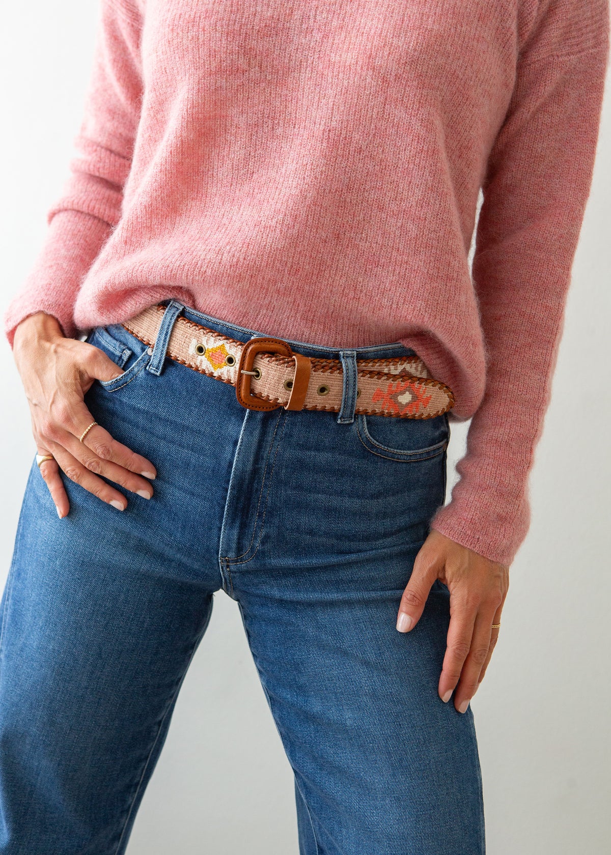 A close-up of a person wearing a pink knit sweater, blue jeans, and a decorative Guatemalan belt with a brown leather buckle. The belt has a tan base with colorful geometric patterns in orange, yellow, and white.