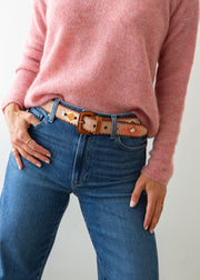 A close-up of a person wearing a pink knit sweater, blue jeans, and a decorative Guatemalan belt with a brown leather buckle. The belt has a tan base with colorful geometric patterns in orange, yellow, and white.