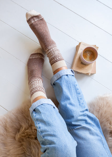 A top-down view of a person's legs wearing brown and cream knit socks with a Fair Isle pattern, paired with light blue jeans, sitting on a fluffy beige rug next to a cup of coffee on a wooden floor.