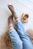 A top-down view of a person's legs wearing brown and cream knit socks with a Fair Isle pattern, paired with light blue jeans, sitting on a fluffy beige rug next to a cup of coffee on a wooden floor.