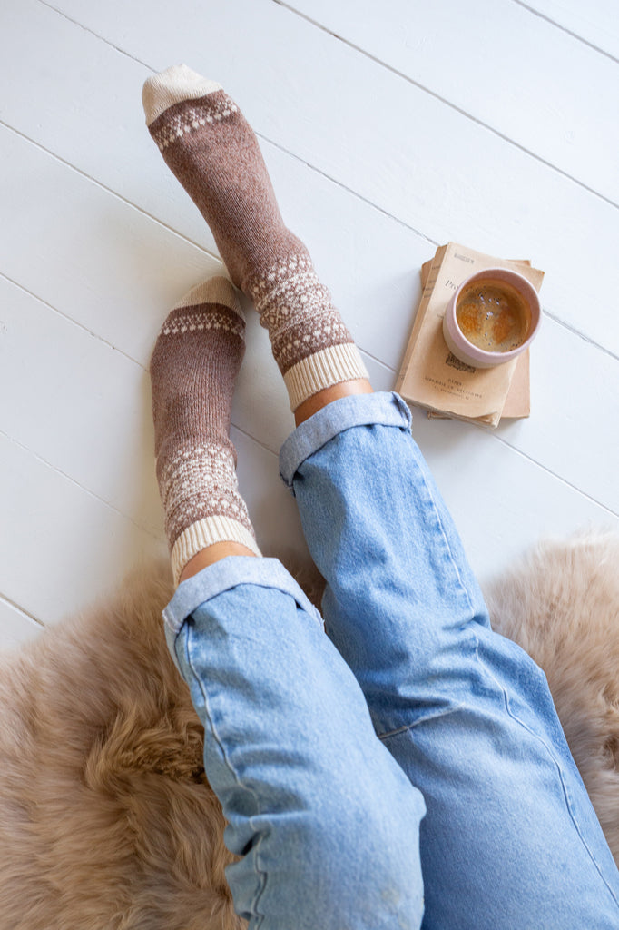 A top-down view of a person's legs wearing brown and cream knit socks with a Fair Isle pattern, paired with light blue jeans, sitting on a fluffy beige rug next to a cup of coffee on a wooden floor.