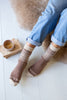 A person wearing brown and cream patterned knit socks with light blue jeans, sitting on a white wooden floor next to a cup of tea and a fluffy rug.