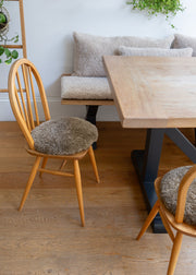 A dining room scene with a light wood table and two wooden chairs with round, textured taupe bouclé seat cushions, set on a warm wood floor in front of a white wall and a matching bench.