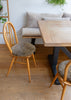 A dining room scene with a light wood table and two wooden chairs with round, textured taupe bouclé seat cushions, set on a warm wood floor in front of a white wall and a matching bench.