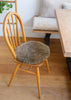 A round, shaggy brown cushion resting on a light wood spindle-back chair in a bright room with hardwood floors and a wooden table.