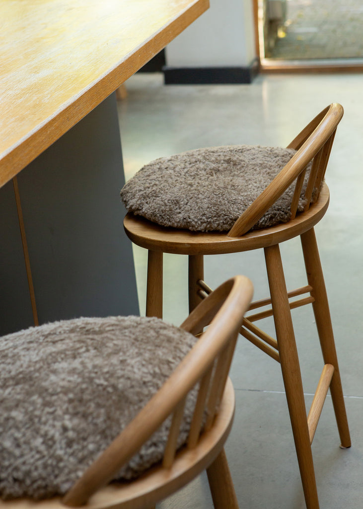 Two wooden bar stools with round, shaggy taupe-gray cushions, positioned next to a light wood counter in a modern interior setting.