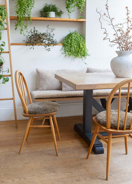 A modern dining area featuring a light wood table with a black trestle base, surrounded by two wooden spindle-back chairs with textured gray bouclé cushions. A built-in wooden bench with matching cushions is set against a white wall with open wooden shelves holding green plants.