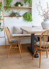 A modern dining area featuring a light wood table with a black trestle base, surrounded by two wooden spindle-back chairs with textured gray bouclé cushions. A built-in wooden bench with matching cushions is set against a white wall with open wooden shelves holding green plants.