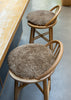 Two round, shaggy taupe-brown bar stools with light natural wood frames, positioned next to a wooden counter on a gray concrete floor.