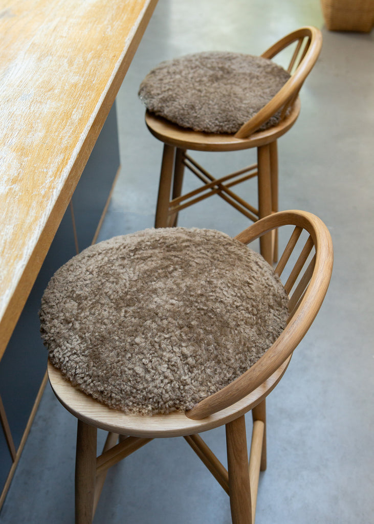Two round, shaggy taupe-brown bar stools with light natural wood frames, positioned next to a wooden counter on a gray concrete floor.