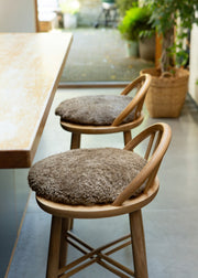 Two wooden bar stools with round, shaggy taupe-brown seat cushions, positioned next to a light wood table in a bright, modern interior.