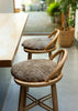 Two wooden bar stools with round, shaggy taupe-brown seat cushions, positioned next to a light wood table in a bright, modern interior.
