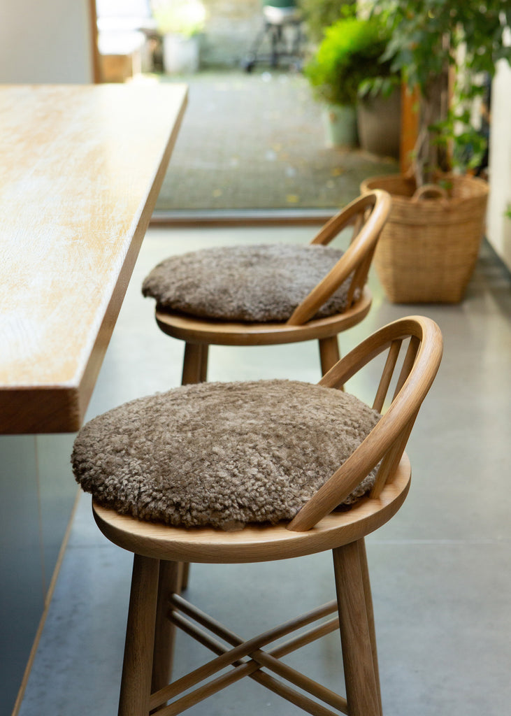 Two wooden bar stools with round, shaggy taupe-brown seat cushions, positioned next to a light wood table in a bright, modern interior.