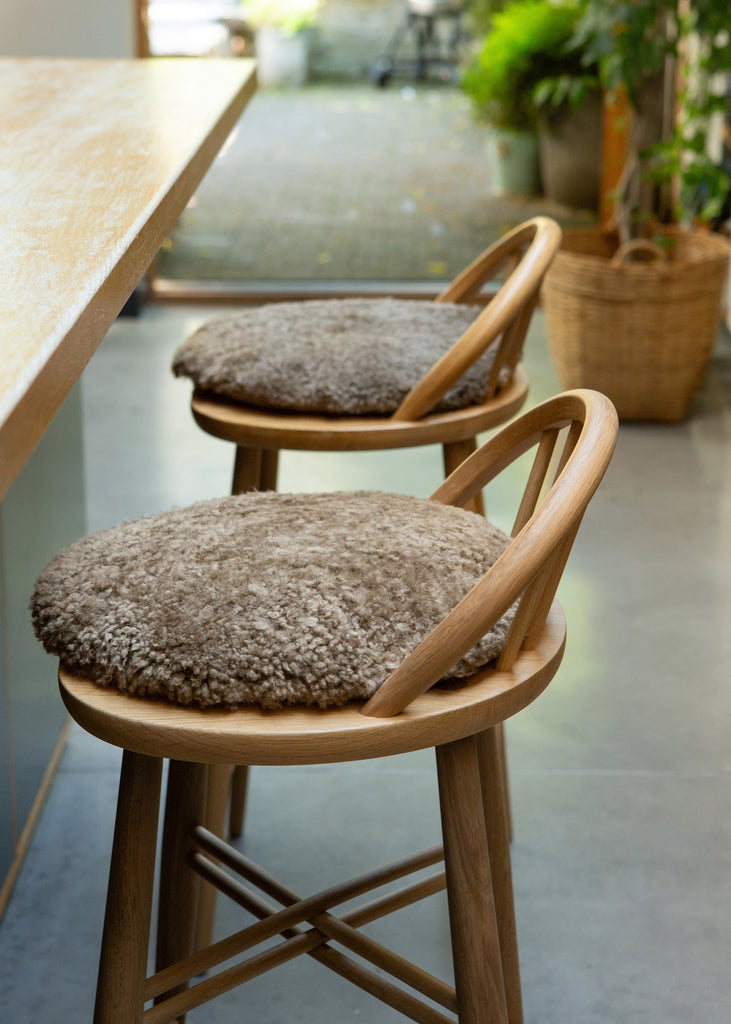 Two wooden bar stools with round, textured taupe boucle cushions, positioned in a modern interior setting next to a light wood counter.