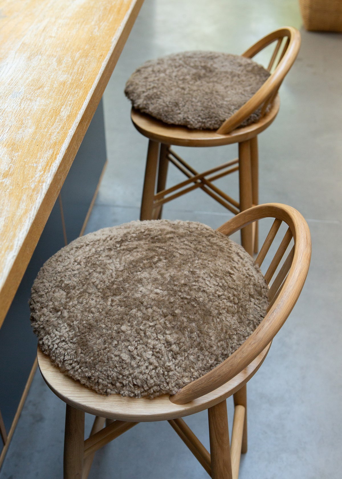 Two round, shaggy brown bar stools with light wood frames and thick, textured brown seat cushions, positioned next to a wooden counter on a gray floor.