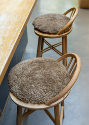 Two round, shaggy brown bar stools with light wood frames and thick, textured brown seat cushions, positioned next to a wooden counter on a gray floor.