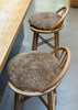 Two round, shaggy brown bar stools with light wood frames and thick, textured brown seat cushions, positioned next to a wooden counter on a gray floor.