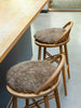 Two wooden bar stools with round, shaggy taupe-brown seat cushions, positioned next to a light wood counter in a modern interior setting.