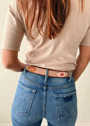 A close-up of a person wearing a pink knit sweater, blue jeans, and a decorative Guatemalan belt with a brown leather buckle. The belt has a tan base with colorful geometric patterns in orange, yellow, and white.
