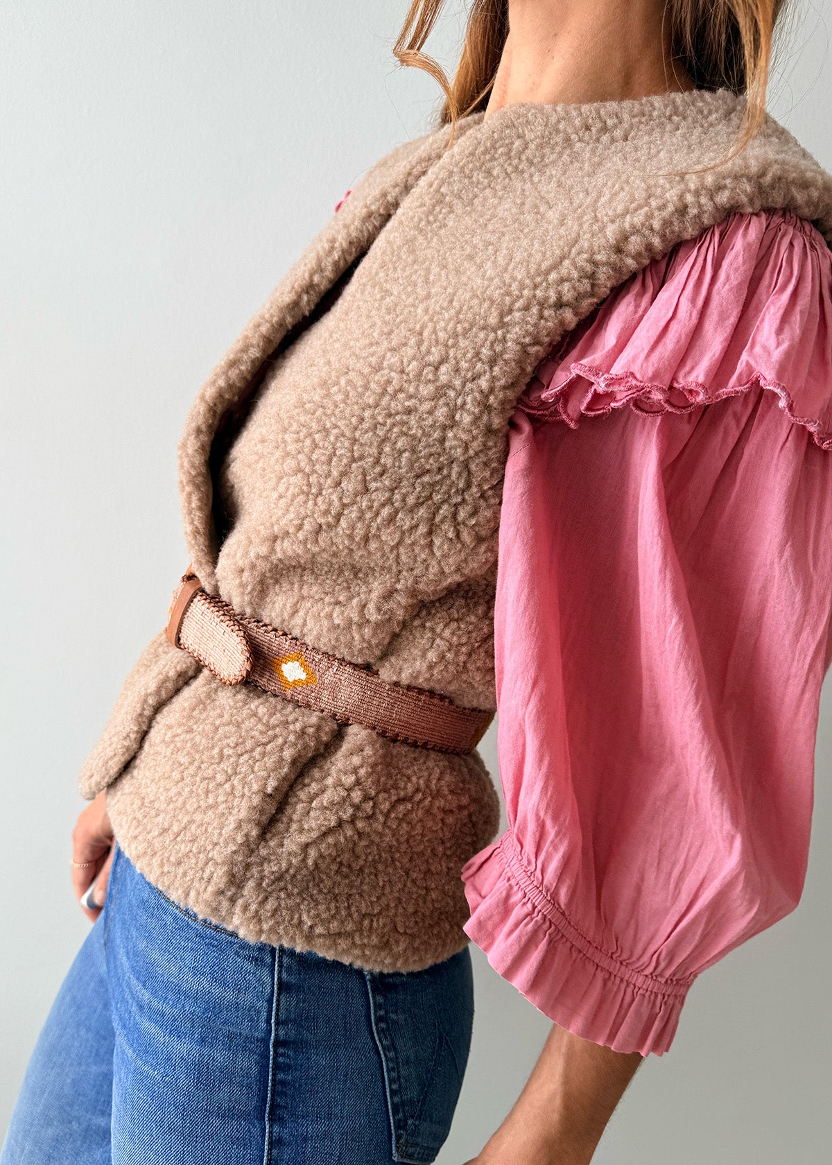 A close-up of a person wearing a pink knit sweater, blue jeans, and a decorative Guatemalan belt with a brown leather buckle. The belt has a tan base with colorful geometric patterns in orange, yellow, and white.