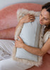 A woman sits on a pink velvet sofa hugging a rectangular light gray pillow with a fluffy cream sheepskin border.