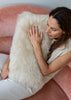 A woman sits on a dusty pink velvet armchair, hugging a fluffy cream sheepskinpillow. She is wearing a cream knit sleeveless top and light-colored pants.