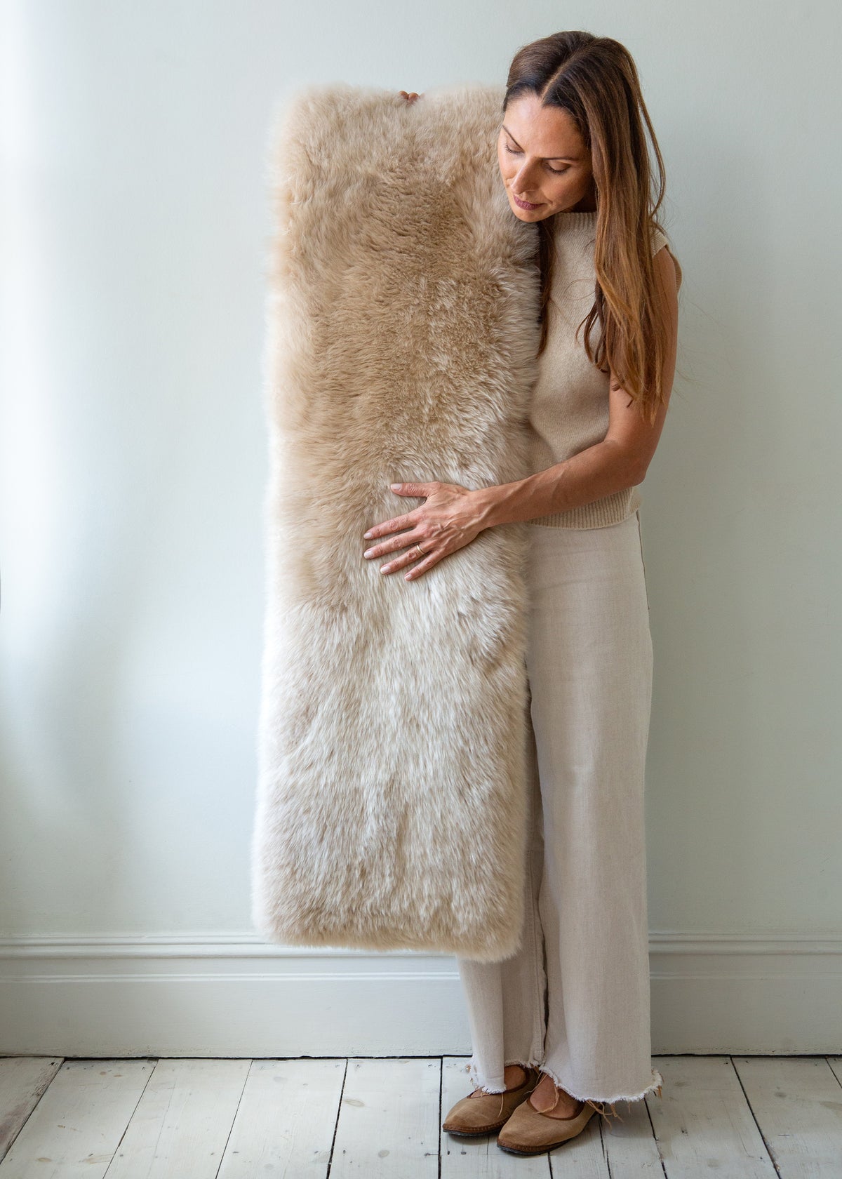 A woman in a beige dress holds a large, fluffy sheepskin bench pad against a light gray wall and white wooden floor.