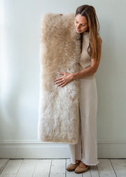 A woman in a beige dress holds a large, fluffy sheepskin bench pad against a light gray wall and white wooden floor.