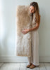 A woman in a beige dress holds a large, fluffy sheepskin bench pad against a light gray wall and white wooden floor.