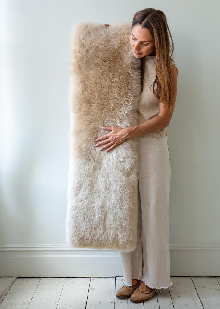 A woman in a beige dress holds a large, fluffy sheepskin bench pad against a light gray wall and white wooden floor.